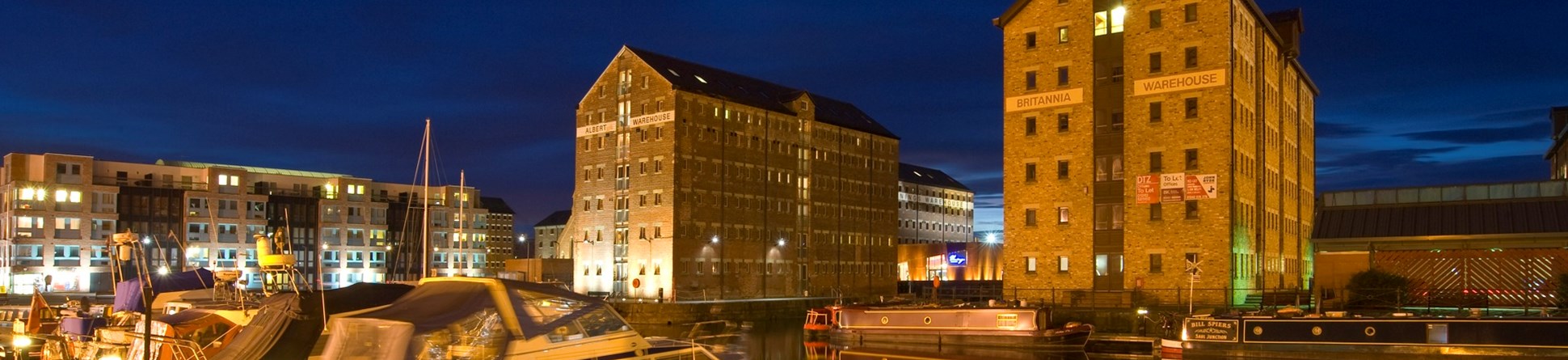 Night-time at Gloucester docks. The street lights pick out boats in the foreground. Converted warehouses stand in the background.