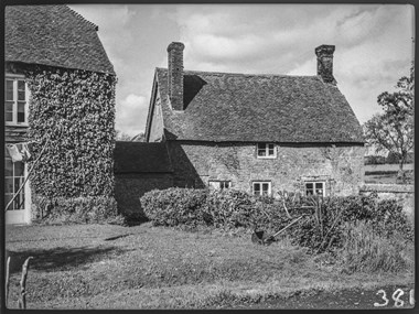 Black and white photograph showing part of a two-storey house clad in foliage and a section of its garden, and, to the side, a stone-built cottage with pitched roof and brick chimney stacks. To the left of the garden is a washing line with some garments pegged to it, and at the edge of the garden lawn is a small, manual lawn mower.