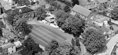 This 1928 black and white aerial image shows a large building, Paston Grammar School, to the right of the image with playing fields stretching away from it, surrounded by trees. There are houses and other buildings that surround the school site.
