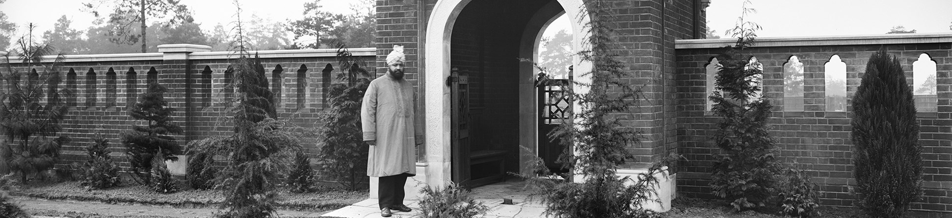 A man wearing a turban stands in front of the Indian inspired gateway of the Muslim Burial Ground, Horsell Common, Surrey. A wall pierced with Indian style tracery surrounds the cemetery.