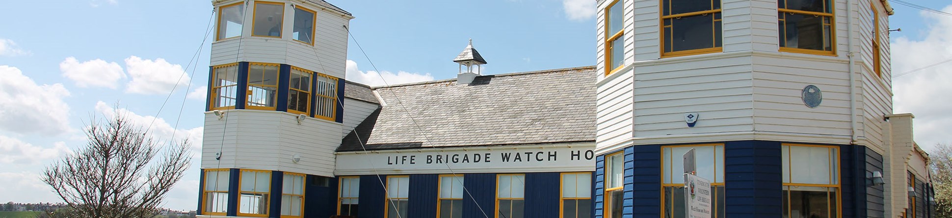 Photo of the outside of the Life Brigade Watch House - a white wooden building with blue and yellow painted windows.