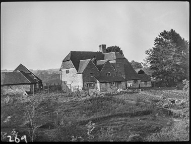 Black and white photograph showing the side and rear elevation of a large farmhouse faced in stone and tile-hanging. Washing hangs outside the back door. To the left of the farmhouse are single-storey farm buildings. In the foreground is a patch of uncultivated ground adjacent to a vegetable garden.
