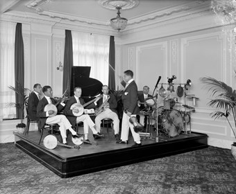 Band playing drums, saxaphone and banjos sitting on a platform with the conductor holding a violin and using the bow as a baton