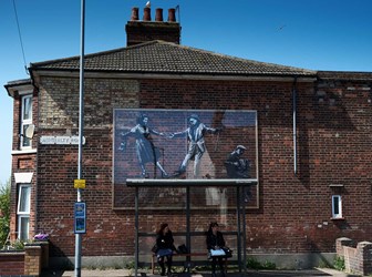 A mural by street artist Banksy above a bus stop depicts a man and woman dancing to music being played on an accordion. Two people are sitting at the bus stop waiting for a bus.
