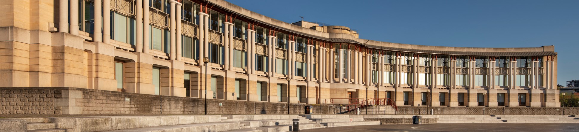 A grand, modern building with a curved frontage and colonnade overlooks a paved public space