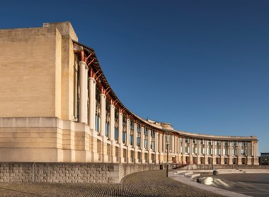 An  exterior view of a long, crescent shaped building. It is two stories high and has paired columns supporting a steel roof structure. In front of it is a paved amphitheatre, or open air space. 