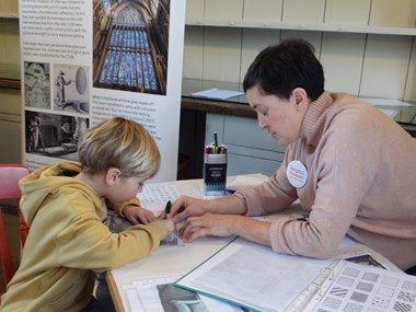 A young boy at a display stand is colouring in a transparent sheet in the style of stained glass, aided by a woman seated opposite.