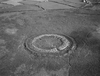 An aerial photograph taken at low altitude showing a circular multivallate hillfort in heathland. Upright stones mark the entrance to the inner enclosure.
