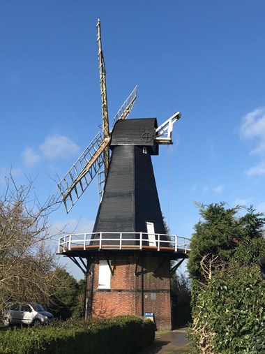Portrait photograph of windmill, with red brick on the lower half and black wooden panels on the top half. The sky is blue and there are green trees and bushes surrounding the windmill.