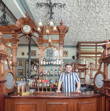 Barman smiling behind the bar, surrounded by decorative woodwork and glass features.