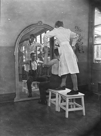 A physiotherapist directing remedial exercises, whilst standing on a stool behind a patient seated in front of a mirror