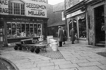 archive black and white photograph of shoppers, wheelbarrows and dustbins displayed on the pavement in front of shops