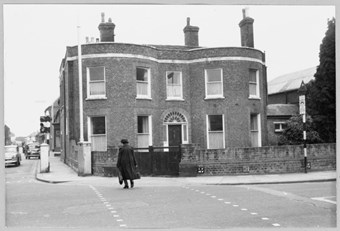 archive black and white photograph of a double-bayed house on a street corner with a pedestrian crossing the road in the foreground