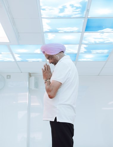 A man in a turban prays in a room with sky-patterned ceiling tiles. 