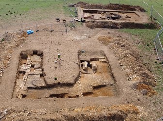An aerial view of two trenches at an archaeological excavation containing stone  foundations of buildings and structures. In the centre is a tripod for surveying equipment.