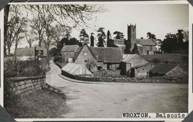 Black and white photograph of a rural street scene featuring empty roads, houses, outbuildings, and a parish church. In the foreground, the road narrows, probably at a junction, with one road continuing into the distance. To the left is a bank of grass, a coursed stone wall with coping and a pub sign reading: 'The Butchers Arms'. To the right of the narrow road are stone houses and outbuildings. A narrow pavement curves around the low wall to the central farmhouse and yard. Behind, and on a slight incline, is a church with a square tower set at one side of the nave. A label applied to the bottom-right of the photograph reads: 'Wroxton. Balscote'. The four corners of the photograph are obscured by photograph album mounts.