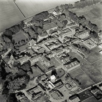 Black and white aerial photo showing a complex of conjoined buildings, several set around courtyards with surrounding lawns