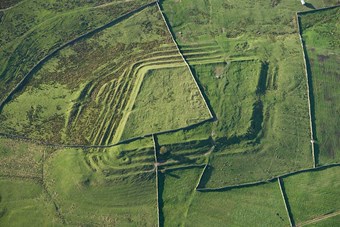 Colour aerial photo showing the multiple banks of a lozenge shaped earthwork cut by dry stone walls and set in moorland