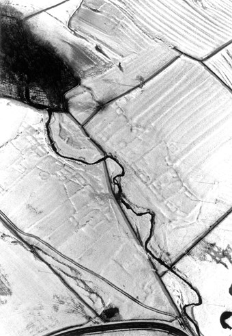 Black and white aerial photo showing low earth banks of enclosures and fields picked out by snow arranged along a stream