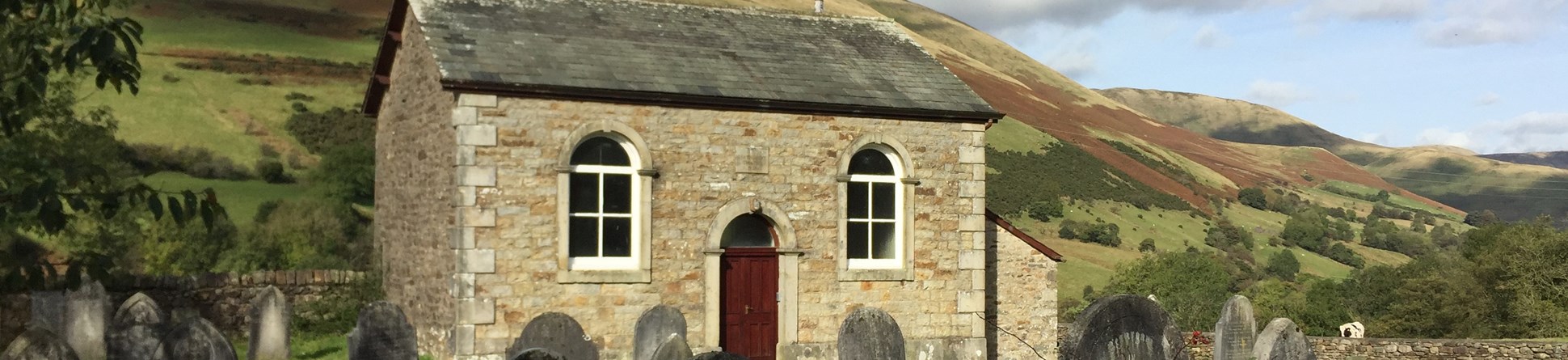 Small stone chapel set in Cumbrian mountain scenery on a sunny day.