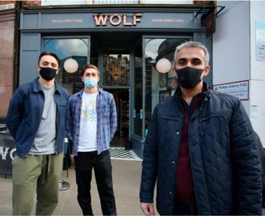 Three men in face masks stand outside a restaurant with a sign saying "Wolf"