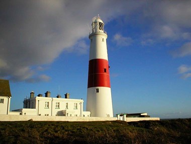 Blue sky with grey clouds with a red and white lighthouse and cottage in the centre of the image and grass in the foreground 