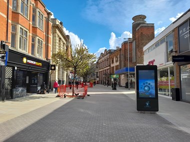 View along a pedestrianised high street with barriers and bollards in place around trees growing out of the paving. In the foreground on the right hand side is an electronic advertisement panel.