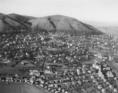 An aerial photograph showing a low-density town situated at the base of a range of undulating hills. Low sunlight illuminates houses in large gardens.
