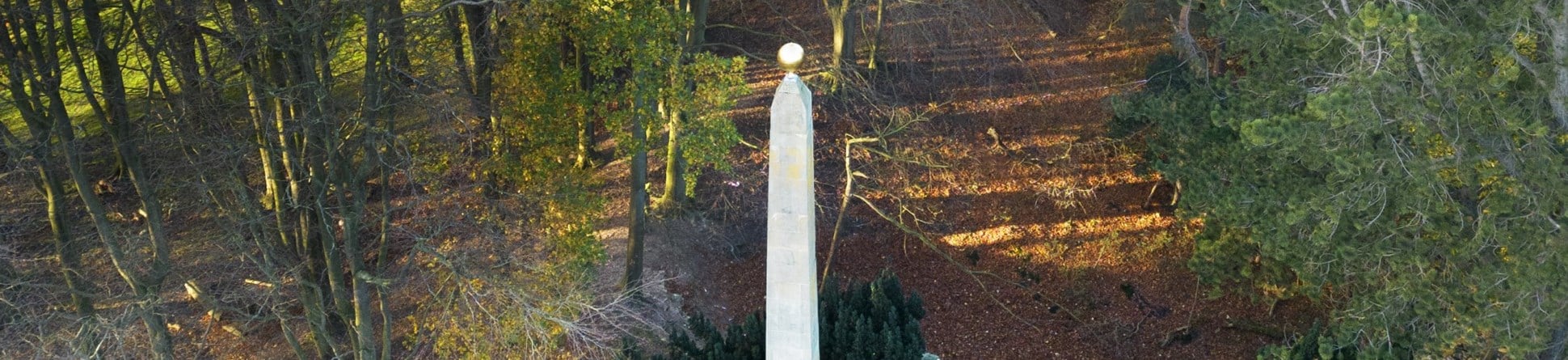 An obelisk monument in a large garden and woodland setting. 