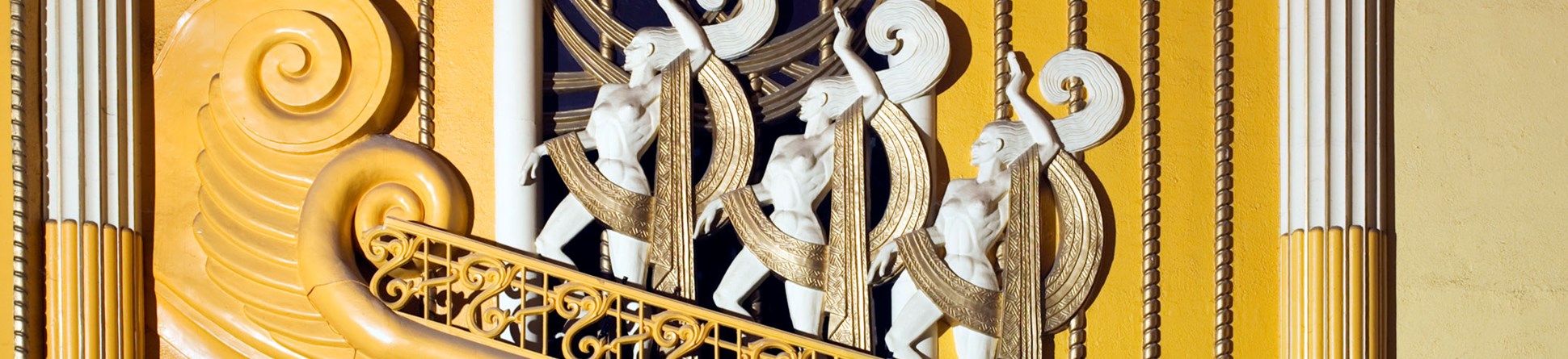 A detail of plaster decoration in vibrant yellow and white, including a row of three mythical figures in an art deco style.