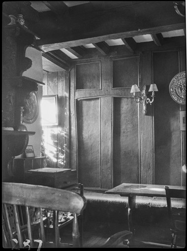 Black and white photograph of a corner of a room in an inn. Timber furniture stands in front of a plank and muntin screen, a section of which features a cross shape. Sunlight enters the room from an obscured opening to the left of the screen. The ceiling features a large timber cross beam and joists.