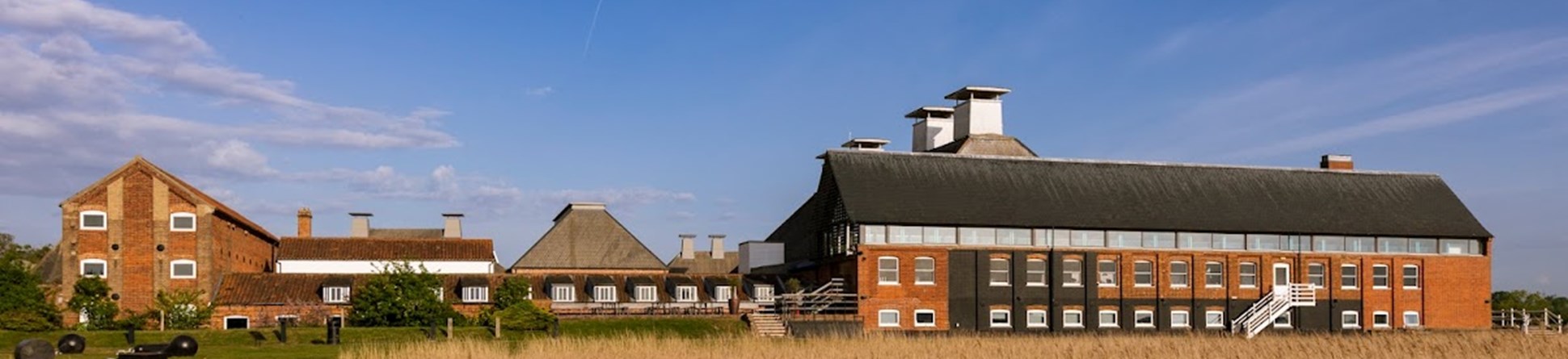 Through wheat fields, in the distance are a collection of red brick buildings, with a large long building to the right, small buildings in the centre and the front of a long building to the left.