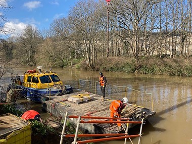 On a river a small boat is moored alongside a pontoon. Building materials have been unloaded on to the pontoon by workmen.