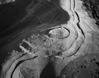 An aerial photograph of an Iron Age hillfort. Strong sunlight casts shadows that reveal banks, ditches and tracks surrounding a circular mound.