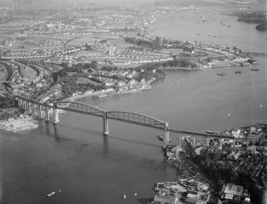 Black and white oblique aerial photograph of a railway bridge spanning a river. The centre of the bridge is dominated by two wide spans carrying massive arched trusses. Boats and barges dot the river. Beyond the bridge are rows of new estate housing and in the far distance are docks harbouring large ships, including two aircraft carriers.