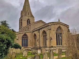 A path runs through a graveyard, flanked by tombstones. A traditional sandstone church stands among them. 