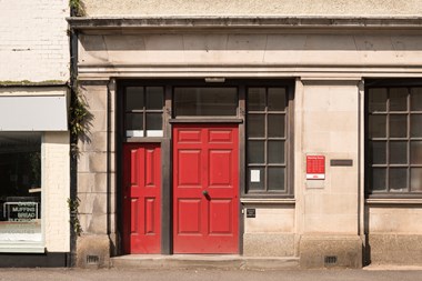 A picture of a stone building with a red, wooden panelled door. It is the front entrance to a historic post office building.