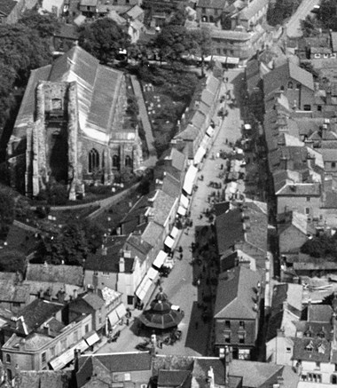 This black and white 1928 aerial photograph shows the market place. It is market day and stalls can be seen extending along the right side of the market place. The church can be seen to the south and the Market Cross can be seen at the front of the image.