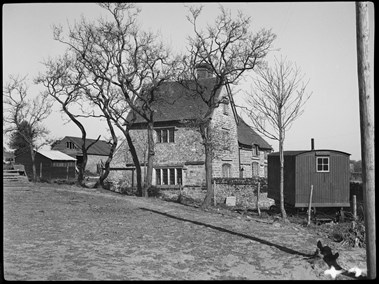 Black and white photograph of a stone-built farmhouse behind a line of bare trees. Taken from across a patch of open ground, the view shows one end of the farmhouse and an oblique view of a long elevation. To one side of the photograph is a shepherd's hut. In the background, to the left of the farmhouse, are farm buildings.