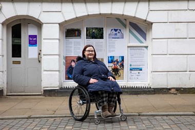 Full-length portrait of a young woman in a wheelchair posing for this photo on the street outside premises.