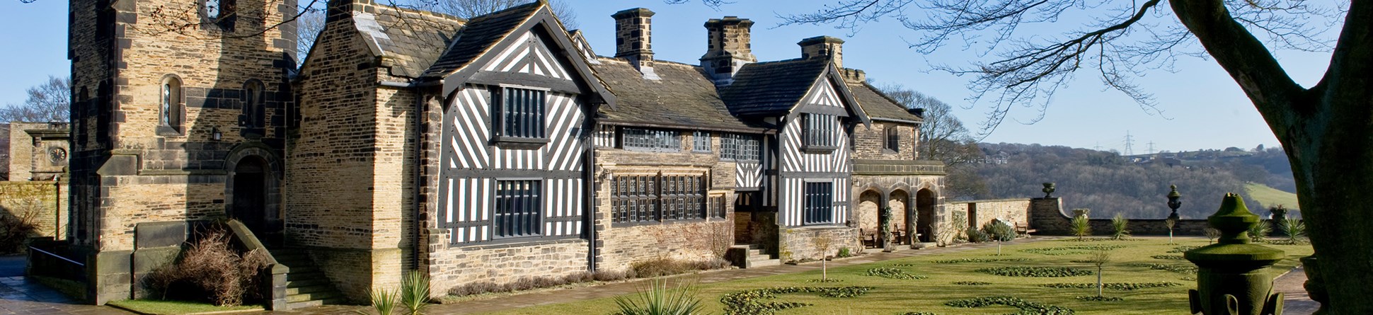 Photo of Shibden Hall, a long house with a mix of timber-framed and stonework walls. A stone tower stands slightly behind the house, closest to the camera. There are manicured gardens before the house and the path is lined with stone urns.