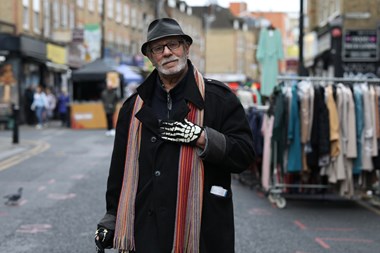 Portrait photo of a man in coat, hat, scarf and gloves, standing on a street with market stalls in the background.