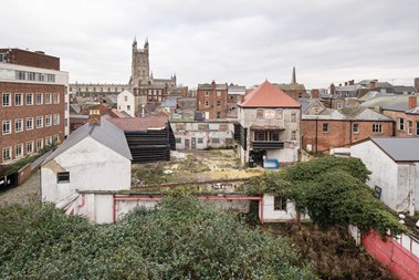 A view across the city of Gloucester with the cathedral in the middle distance. In the foreground is a collection of buildings in poor repair set around a derelict courtyard. A sign on the upper floor of one of the buildings reads 'Fleece Hotel'.