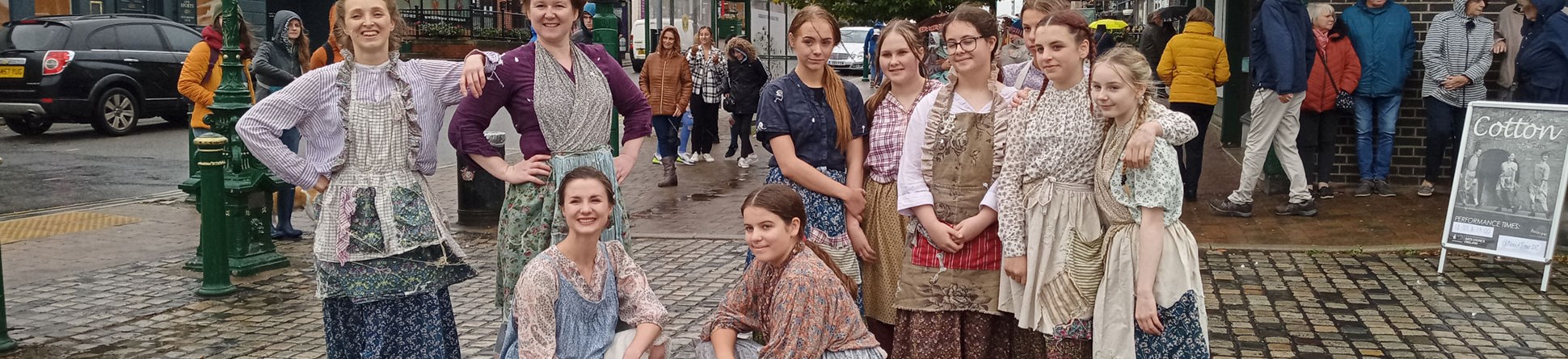 Young women and schoolgirls dressed as Victorian or Edwardian mill girls pose together for a photograph in a pedestrian precinct.