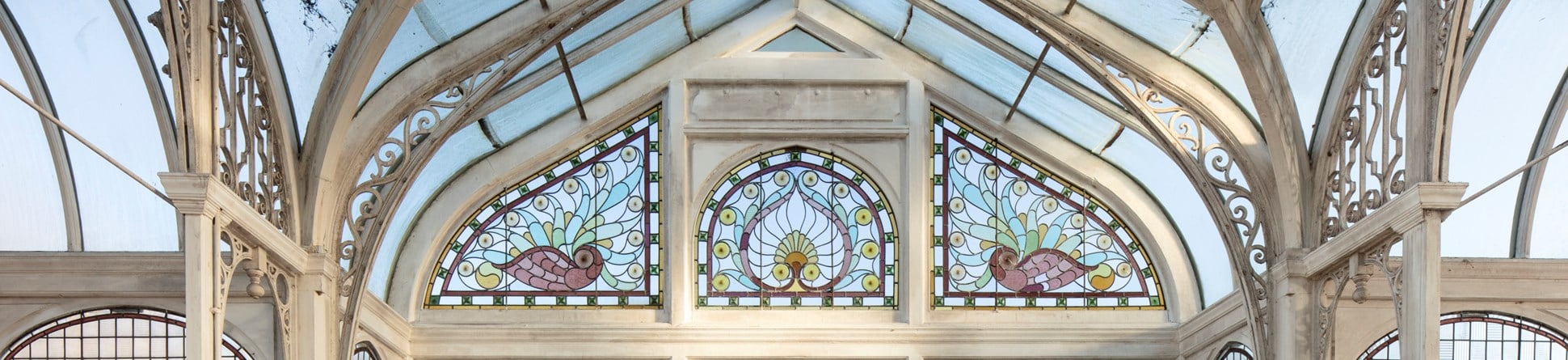 A white framed conservatory with decorative scrolled ironwork and, above plain glass windows, three beautifully decorated panels showing birds and a central peacock feather design.