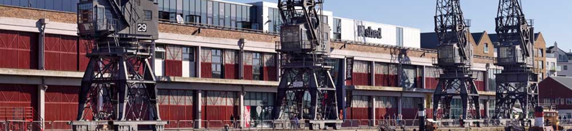 A photograph of four large, grey, 20th century cranes on a wharf with a long industrial building behind them.