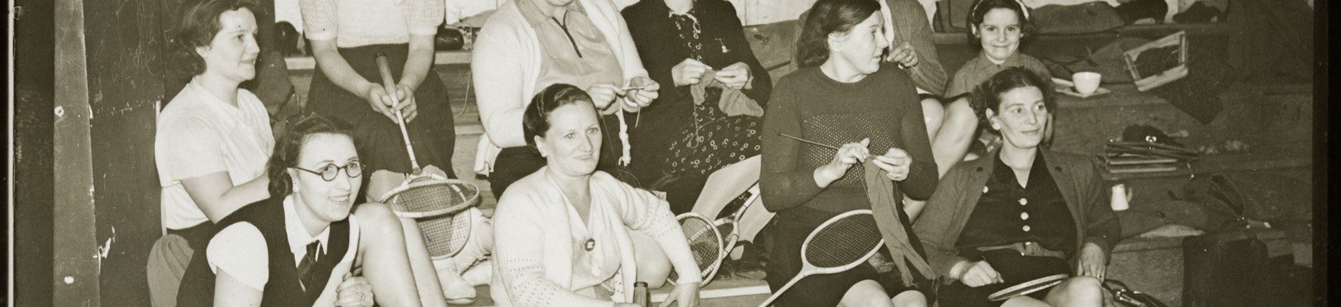 Women sitting on tiered benches holding badminton racquets and knitting.