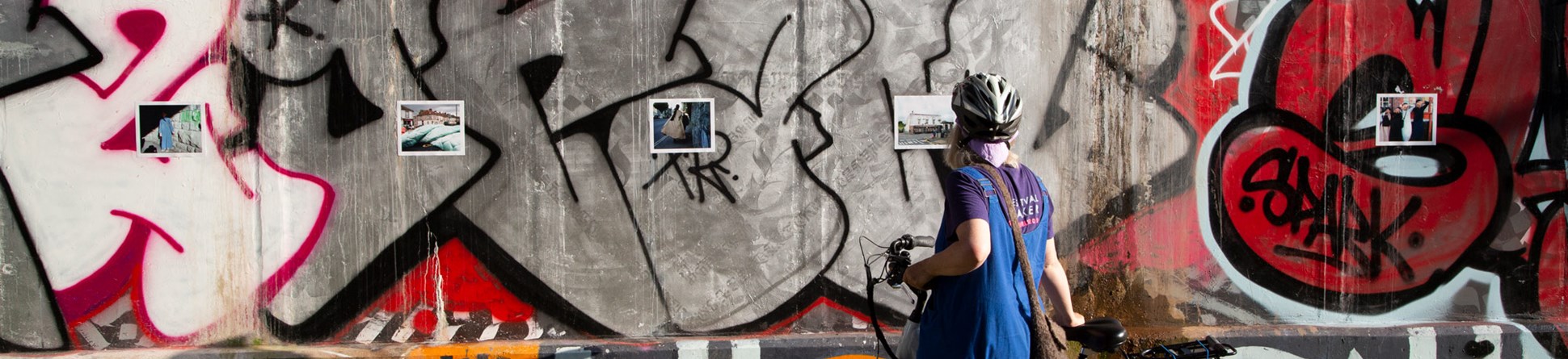 Person standing with bike in front of postcard size photographs mounted on a concrete graffiti wall.