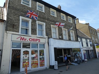 Three-storey brick built building with Mr. Chips chip shop on ground floor. Three flag poles project from the front. One with a Union Jack, one with a St George's cross and one not visible.