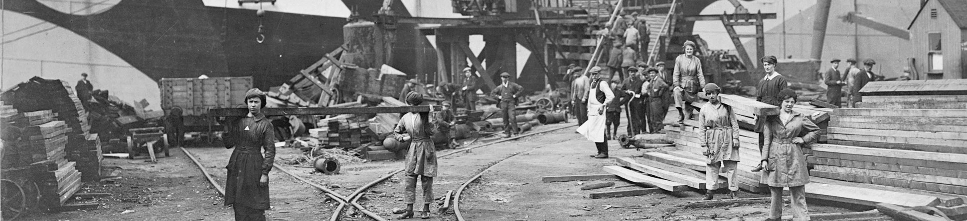 A black and white image of a shipbuilding yard with a boat in the background and women carrying timber. 